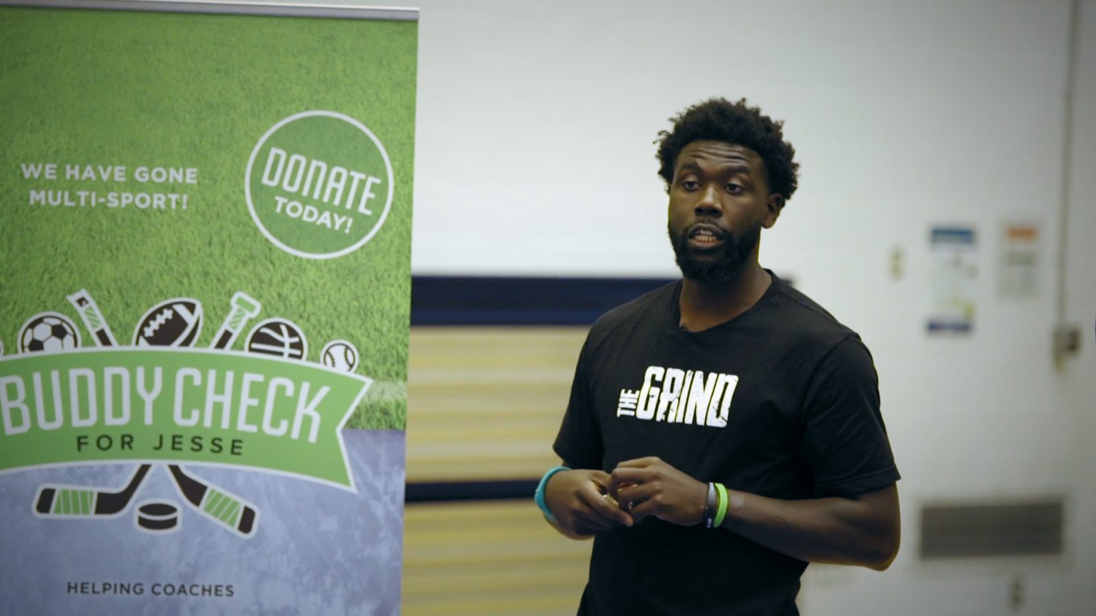 A coach in a 'The Grind' shirt beside a Buddy Check banner
